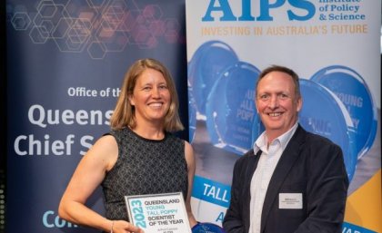 Two people stand smiling in front of banners at an awards night, one of them is holding an award.